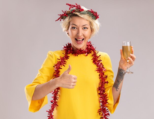 Vrolijke jonge blonde vrouw die de hoofdkroon van Kerstmis en klatergoudslinger om hals draagt die glas champagne houdt op zoek toont duim omhoog geïsoleerd op witte muur