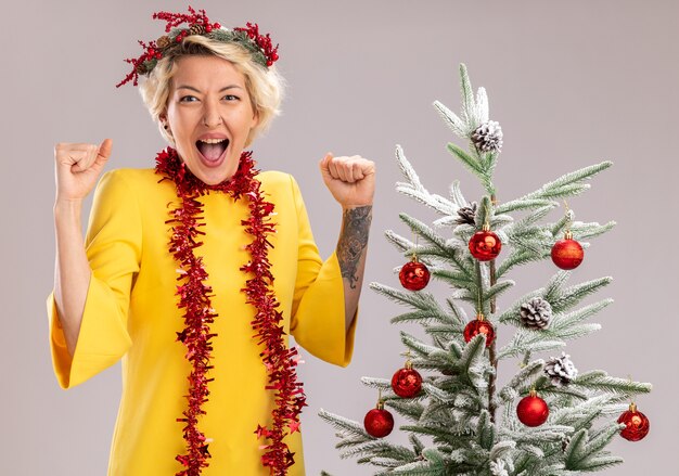 Vrolijke jonge blonde vrouw die de hoofdkroon van Kerstmis en klatergoudslinger om hals draagt die zich dichtbij versierde kerstboom bevindt die op zoek doet ja gebaar geïsoleerd op witte muur