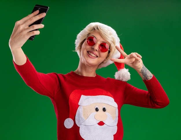 Vrolijke jonge blonde vrouw met kerstmuts en kerstman kerst trui met bril doen vredesteken nemen selfie geïsoleerd op groene achtergrond