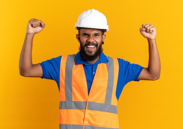 Vrolijke jonge bouwer man in uniform met veiligheidshelm staande met opgeheven vuisten omhoog geïsoleerd op oranje muur met kopieerruimte