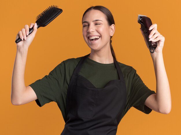 Vrolijke jonge brunette kapper meisje in uniform houdt haartrimmer en kam op oranje