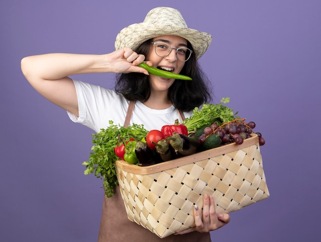 Vrolijke jonge brunette vrouwelijke tuinman in optische bril en in uniform dragen tuinieren hoed houdt groentemand vast en doet alsof hij hete peper bijt geïsoleerd op paarse muur