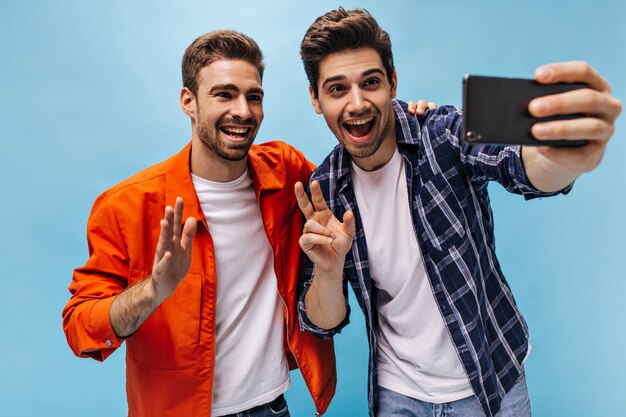 Vrolijke jonge charmante mannen nemen selfie in goed humeur op blauwe achtergrond Cool man in oranje jas golven hand in groet Guy in geruit overhemd houdt telefoon vast en toont vredesteken