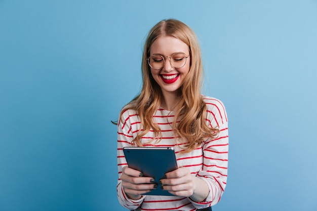 Vrolijke jonge dame die digitale tablet met glimlach houdt. Studio shot van schattige dame in casual kleding geïsoleerd op blauwe achtergrond.