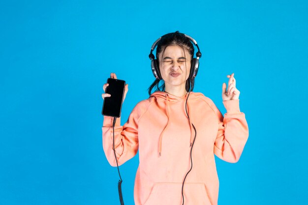 Vrolijke jonge dame die haar telefoon een luistermuziek op blauwe achtergrond houdt