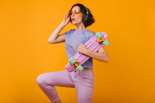 Vrolijke jonge dame in koptelefoon rondkijken terwijl ze poseren op een oranje achtergrond Vrouwelijk model met krullend kapsel met roze skateboard