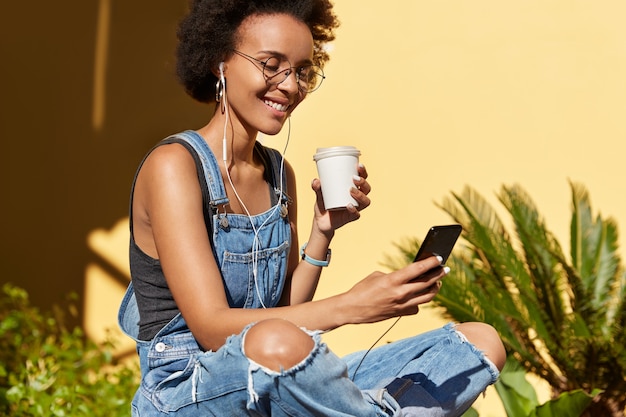 Vrolijke jonge hipster met krullend, borstelig haar, draagt een bril en haveloze overall, downloadt liedjes naar afspeellijst op mobiele telefoon, drinkt een vers drankje uit een wegwerpbeker, geniet van vrije tijd in de zomer