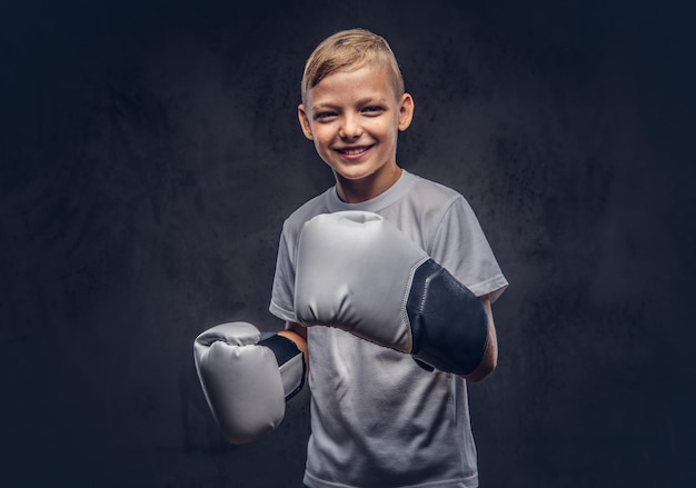 Vrolijke jonge jongen bokser met blond haar gekleed in een wit t-shirt met bokshandschoenen poseren in een studio. Geïsoleerd op de donkere gestructureerde achtergrond.