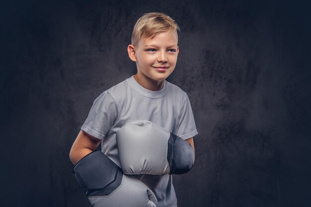 Vrolijke jonge jongen bokser met blond haar gekleed in een wit t-shirt met bokshandschoenen poseren in een studio. Geïsoleerd op de donkere gestructureerde achtergrond.