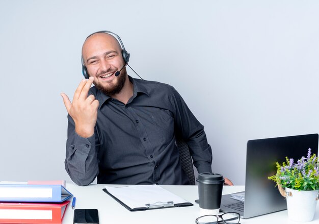 Vrolijke jonge kale callcentermens die hoofdtelefoonszitting bij bureau met uitrustingsstukken draagt ?? die pistoolteken met hand op wit wordt geïsoleerd