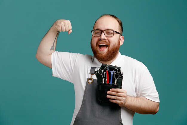 Vrolijke jonge kapper in uniform en bril met kapperstas met kappershulpmiddelen kijkend naar camera met sterk gebaar geïsoleerd op blauwe achtergrond