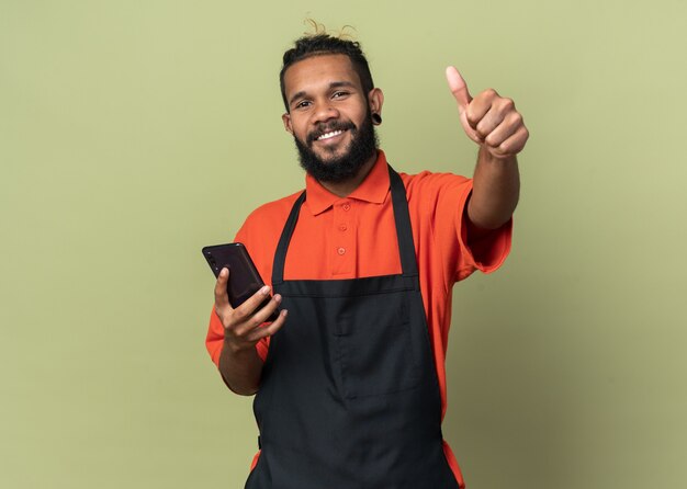 Vrolijke jonge kapper in uniform kijkend naar de voorkant met mobiele telefoon met duim omhoog geïsoleerd op olijfgroene muur met kopieerruimte