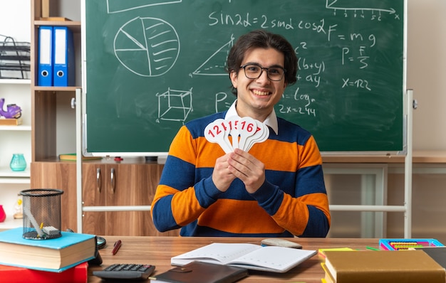 vrolijke, jonge, kaukasische, meetkundeleraar met een bril die aan het bureau zit met schoolbenodigdheden in de klas met nummerfans die naar voren kijken