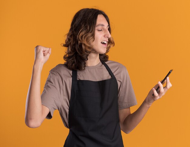 Vrolijke jonge knappe kapper die uniform vasthoudt en naar een mobiele telefoon kijkt die ja gebaar doet