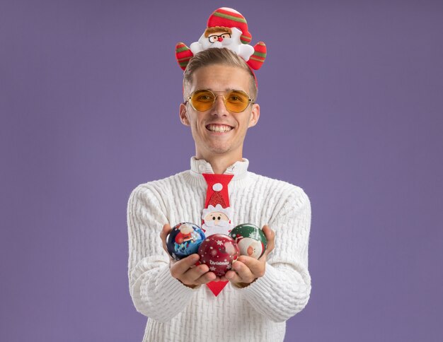 Vrolijke jonge knappe kerel die de hoofdband en de stropdas van de Kerstman met glazen draagt die de ornamenten van de Kerstmisbal houden die op purpere muur met exemplaarruimte worden geïsoleerd