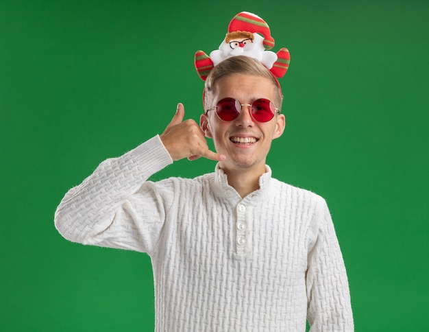 Vrolijke jonge knappe kerel die de hoofdband van de Kerstman met glazen draagt ?? die vraaggebaar doet dat op groene muur wordt geïsoleerd