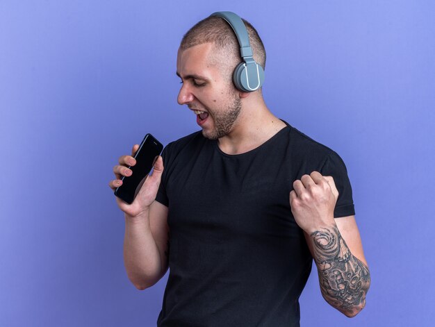 Vrolijke jonge knappe kerel met een zwart t-shirt en koptelefoon met telefoon en zingt geïsoleerd op een blauwe muur