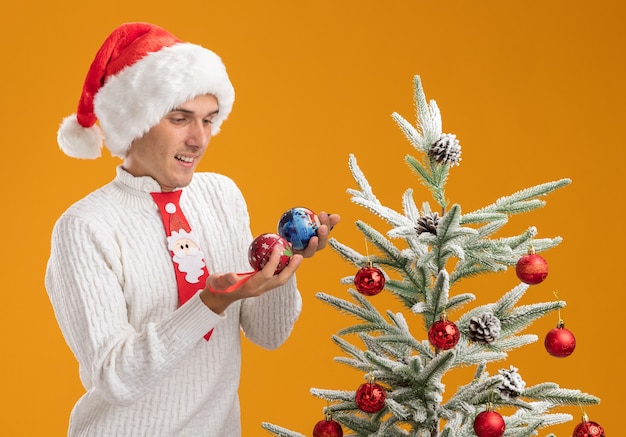 Vrolijke jonge knappe kerel met kerstmuts en stropdas van de kerstman permanent in de buurt van versierde kerstboom houden en kijken naar kerstbal ornamenten geïsoleerd op een oranje achtergrond