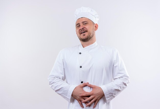 Vrolijke jonge knappe kok in chef-kok uniform handen op buik op geïsoleerde witte muur zetten
