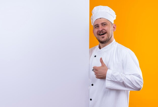 Vrolijke jonge knappe kok in uniform van de chef-kok die achter een witte muur staat en duim omhoog laat zien, geïsoleerd op een oranje muur met kopieerruimte