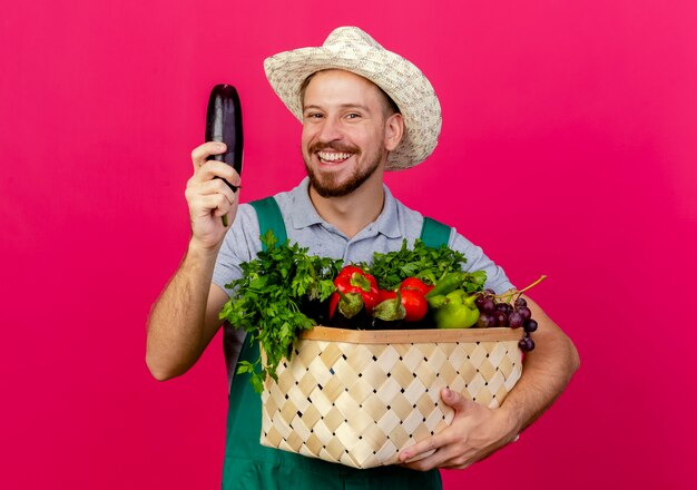 Vrolijke jonge knappe Slavische tuinman in uniform en hoed met mand met groenten en aubergine geïsoleerd op karmozijnrode muur