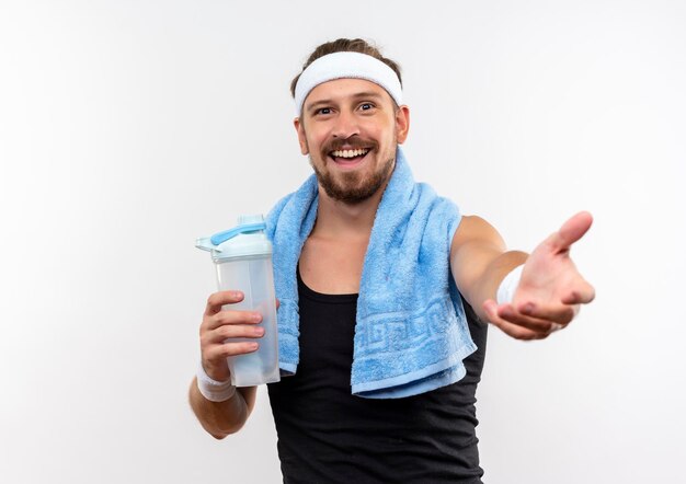 Vrolijke jonge knappe sportieve man met hoofdband en polsbandjes die een waterfles vasthouden en de hand uitstrekken die op een witte muur is geïsoleerd