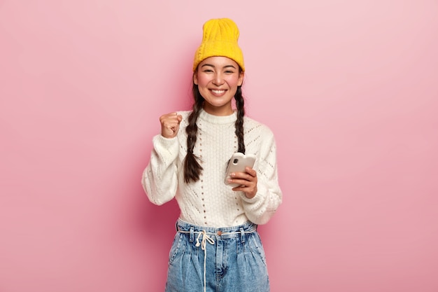 Vrolijke jonge koreaanse vrouw balt vuist, draagt gele stijlvolle hoed en witte trui, glimlacht aangenaam, houdt moderne mobiele telefoon, geïsoleerd over roze studiomuur