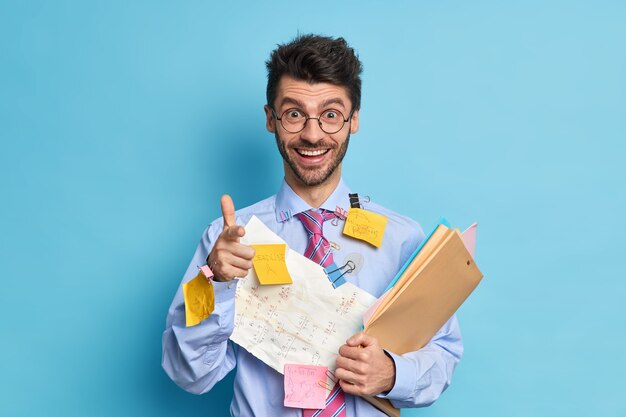 Vrolijke jonge man collega blij om projectwerk bedekt met papieren en stickers af te ronden wijst naar je vinger pistool gebaar maakt. Succesvolle ijverige student bezig met cursuswerk vormt binnen