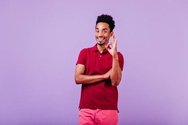 Vrolijke jonge man draagt een rood t-shirt poseren met goed teken. grappig Afrikaans mannelijk model geïsoleerd.