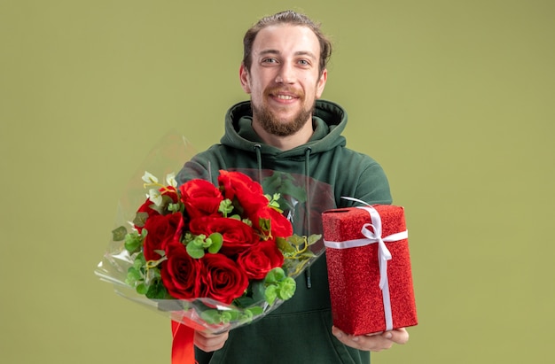 vrolijke, jonge man, in, vrijetijdskleding, met, bouquetten, van, rode rozen, en, cadeau, voor, zijn, vriendin, kijkend naar de camera, het glimlachen, vrolijk, staand, op, groene muur, valentijnsdag, concept