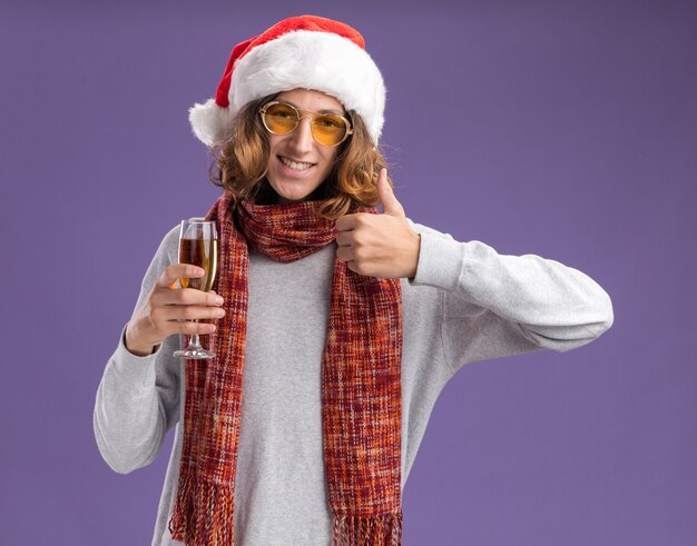 Vrolijke, jonge man, met, kerstmuts, en, gele bril, met, warme sjaal om zijn, nek, vasthouden, glas champagne, kijken naar, camera, het glimlachen, weergeven, beduimelt omhoog, staand, op, paarse achtergrond