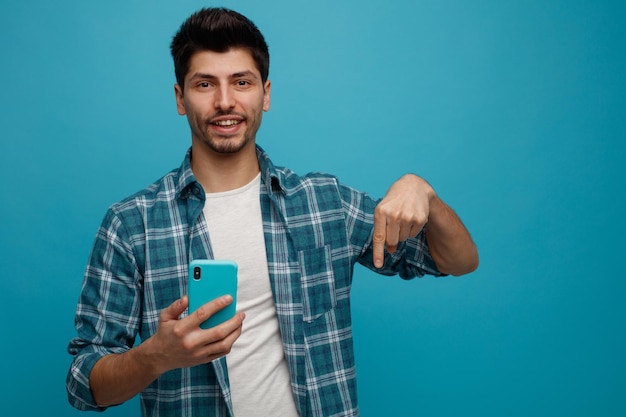 Vrolijke jonge man met mobiele telefoon kijken camera naar beneden geïsoleerd op blauwe achtergrond