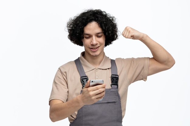 Vrolijke jonge mannelijke bouwvakker die uniform vasthoudt en naar mobiele telefoon kijkt met een sterk gebaar dat op een witte achtergrond wordt geïsoleerd