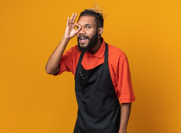 Vrolijke jonge mannelijke kapper met uniform staand in profielweergave kijkend naar de voorkant en doet een gebaar geïsoleerd op een oranje muur met kopieerruimte