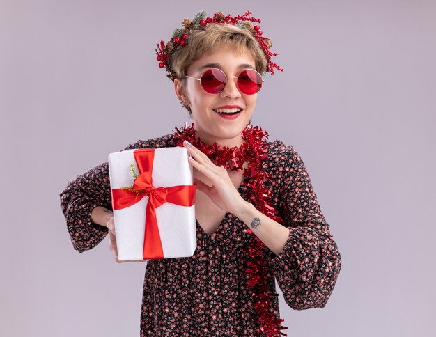 Vrolijke jonge mooi meisje dragen hoofd krans van Kerstmis en klatergoud garland rond nek met bril houden geschenkpakket kijken camera geïsoleerd op witte achtergrond