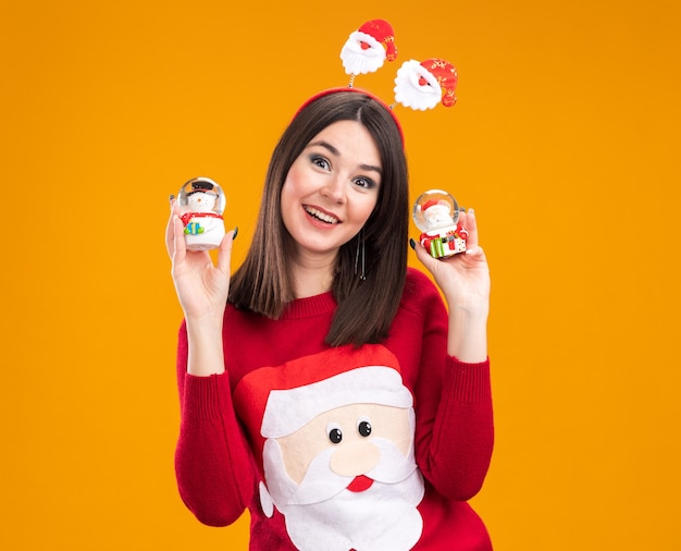 Vrolijke jonge mooie blanke meisje dragen santa claus hoofdband en trui houden sneeuwpop en santa claus beeldjes kijken camera geïsoleerd op oranje achtergrond met kopie ruimte