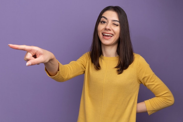 Vrolijke jonge, mooie blanke vrouw die de hand achter de rug houdt en naar de zijkant wijst en knipoogt