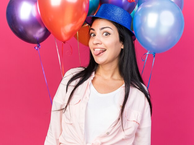 Vrolijke jonge mooie meid met feesthoed die vooraan staat met ballonnen die tong tonen