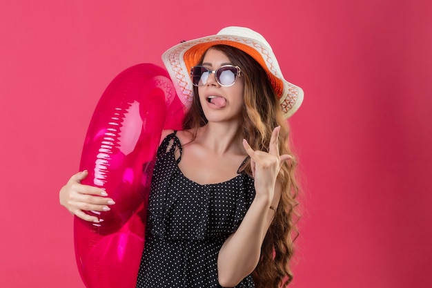 Vrolijke jonge mooie reiziger meisje in jurk in polka dot in zomer hoed zonnebril houden opblaasbare ring blij en positief tong uitsteekt overwinning teken staande over pi maken