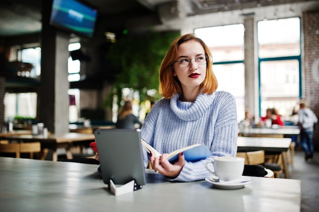 Vrolijke jonge mooie roodharige vrouw in glazen met behulp van haar telefoon touchpad en notebook zittend op haar werkplek op café met kopje koffie