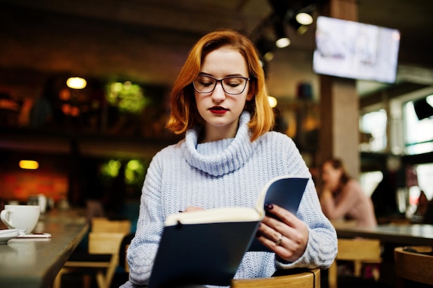 Vrolijke jonge, mooie roodharige vrouw met een bril die op haar werkplek in een café zit en iets leest in haar notitieboekje