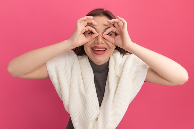 Vrolijke jonge mooie vrouw die een gebaar doet met handen als verrekijker geïsoleerd op roze muur