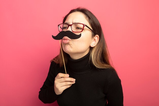 Vrolijke jonge mooie vrouw in een zwarte coltrui en glazen met grappige snor op stok die een kus blaast die zich over roze muur bevindt