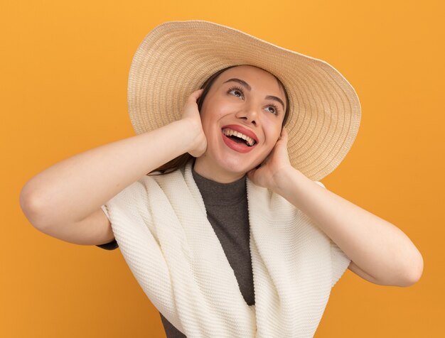 Vrolijke jonge mooie vrouw met een strandhoed die haar handen op het gezicht houdt en omhoog kijkt geïsoleerd op een oranje muur