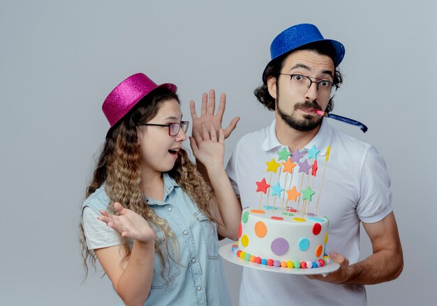 Vrolijke jonge paar dragen roze en blauwe hoeden kerel geeft verjaardagstaart aan meisje en fluitje blazen geïsoleerd op een witte achtergrond