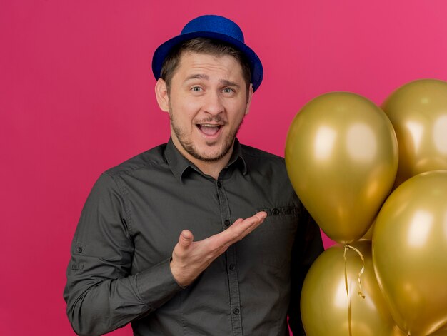 Vrolijke jonge partijkerel die blauwe hoedenholding draagt en wijst met hand op ballons die op roze worden geïsoleerd