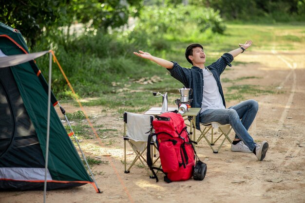 Vrolijke jonge reiziger man zit aan de voorkant van de tent in het bos met koffieset en het maken van verse koffiemolen tijdens het kamperen op zomervakantie