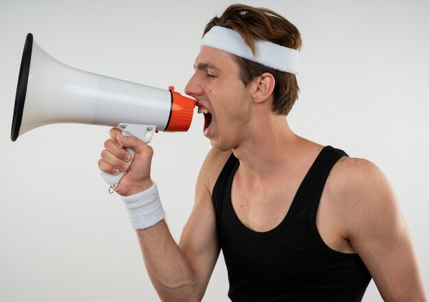 Vrolijke jonge sportieve man met hoofdband en polsbandje spreekt op luidspreker geïsoleerd op een witte muur