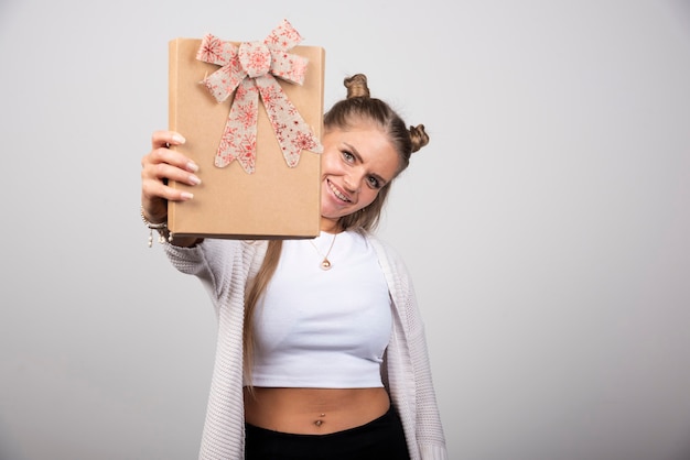 Vrolijke jonge vrouw die geschenkdoos op grijze muur toont.