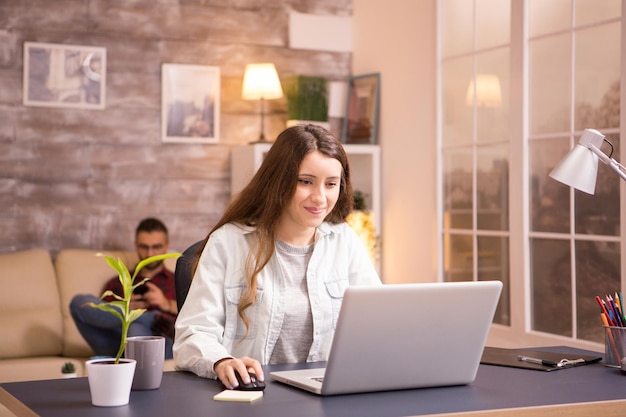 Vrolijke jonge vrouw die lacht tijdens het werken op laptop vanuit huis. Vriendje ontspannen op de bank op de achtergrond.